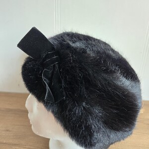 Peut inclure: Un chapeau en fourrure noire avec un nœud en velours. Le chapeau est rond et a un aspect doux et texturé. Le nœud est positionné sur le côté, ajoutant une touche d'élégance. Le chapeau est présenté sur une tête de mannequin blanche.