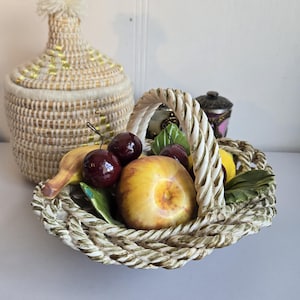 Peut inclure: Un panier en céramique débordant de faux fruits, dont des cerises, une pêche et une banane. Le panier a un design en forme de corde et une poignée blanche. Un pot tissé avec un couvercle et un petit récipient décoratif sont en arrière-plan.