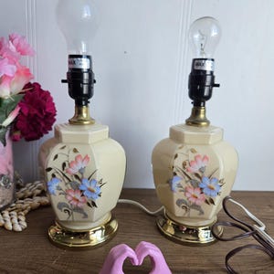 Peut inclure: Paire de lampes de table vintage en céramique avec des motifs floraux. Les lampes ont une base crème avec des accents dorés et présentent un motif floral dans des tons de rose, bleu et vert. Chaque lampe a une douille noire et une ampoule transparente.
