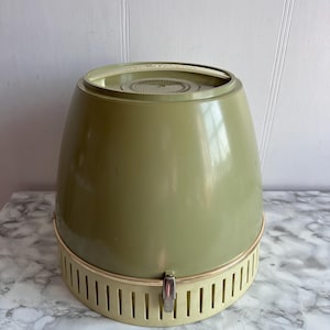May include: Vintage avocado green and cream-colored hair dryer. The hair dryer has a rounded, conical shape with a cream-colored base featuring vertical slots. A silver-colored clasp secures the base. The top is a lighter shade of green.