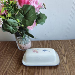 Peut inclure: Un beurrier en céramique blanche avec un motif floral rose, bleu et vert. Le beurrier a une bordure bleue et repose sur un plateau assorti.