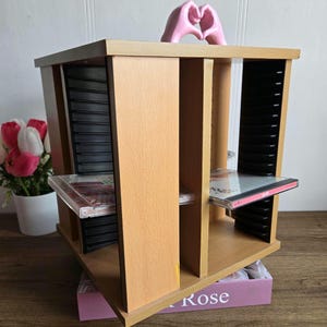 Może przedstawiać: Drewniany schowek na płyty CD w kolorze jasnobrązowym z czarnymi przegrodami, mieszczący płyty CD. Na górze znajduje się różowa rzeźba dłoni tworzących kształt serca. Urządzenie stoi na drewnianej powierzchni.