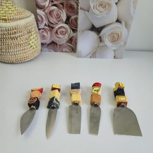 Peut inclure: Ensemble de cinq couteaux à fromage. Chaque couteau est doté d'une lame en acier inoxydable et d'un manche orné de fromages empilés et d'éléments décoratifs. Les manches présentent une variété de couleurs et de textures, ajoutant un intérêt visuel à l'ensemble.