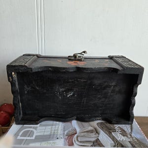 May include: A black wooden chest with a metal clasp and floral accents. The chest is rectangular with a slightly curved top and sits on a stack of books with the words "Home Decor" visible.