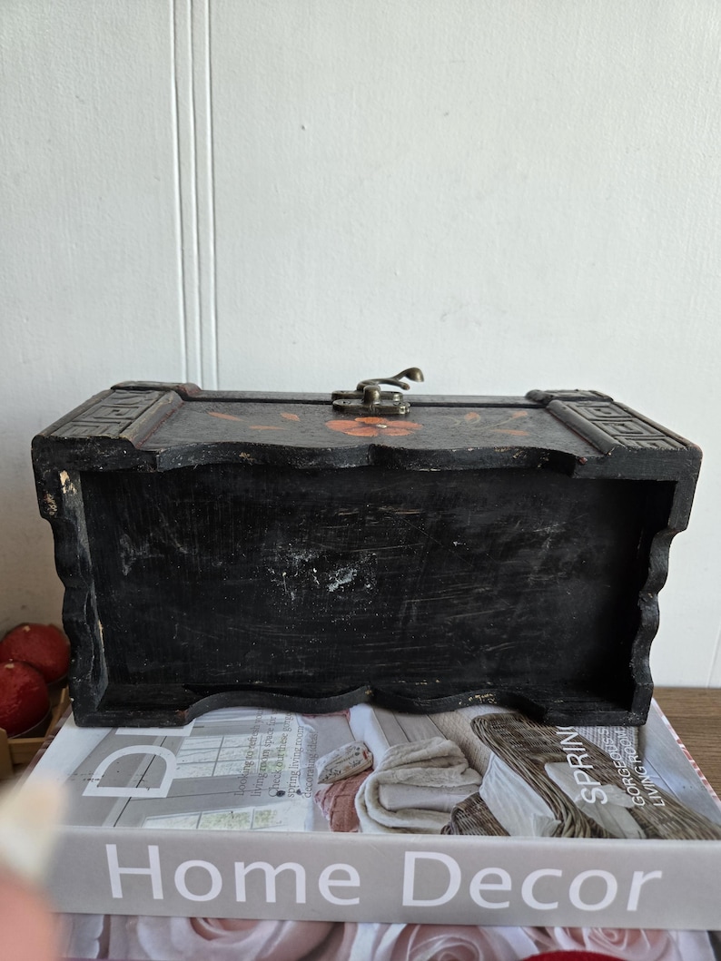 May include: A black wooden chest with a metal clasp and floral accents. The chest is rectangular with a slightly curved top and sits on a stack of books with the words "Home Decor" visible.
