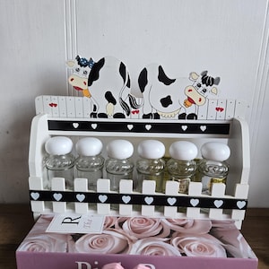 May include: A white wooden spice rack with six glass jars, each with a white lid. The rack features black and white cow figures and a picket fence design. The rack is placed on a box with the words "Pink Rose".