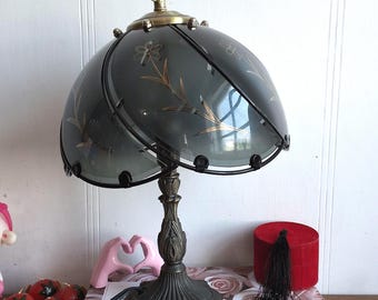 May include: A vintage table lamp with a dark gray glass shade featuring gold floral designs. The lamp has a bronze-colored base and a decorative stem. The lamp is sitting on a book titled "Pink Rose".
