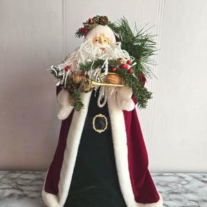 Peut inclure: Un ornement de sommet d'arbre de Noël en velours rouge représentant un Père Noël tenant une trompette dorée et une guirlande verte avec des baies rouges. Le Père Noël a une barbe blanche et une bordure de fourrure blanche sur sa robe.