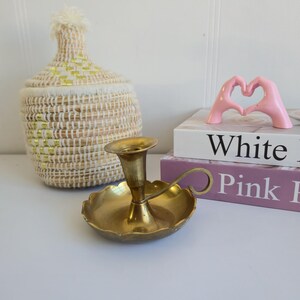 Puede incluir: Un candelero de latón con base y asa festoneadas se encuentra sobre una superficie blanca. También hay una cesta tejida con tapa y una escultura rosa con forma de mano. La palabra "White" es visible en un libro.