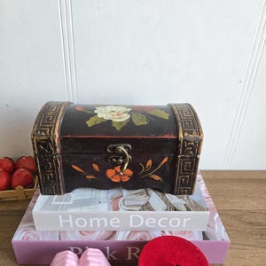 May include: A small, dark wooden chest with floral artwork and gold accents. The chest is placed on top of a stack of books, including one that reads "Home Decor". A pink hand sculpture forms a heart shape in front of the books.