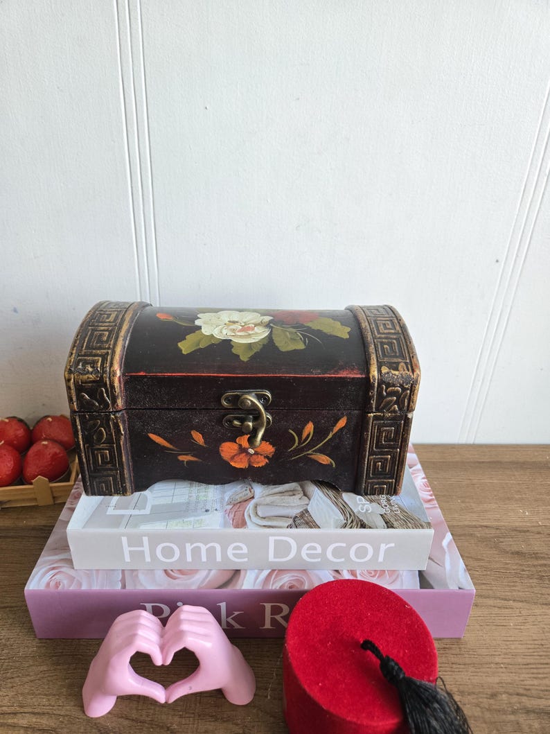 May include: A small, dark wooden chest with floral artwork and gold accents. The chest is placed on top of a stack of books, including one that reads "Home Decor". A pink hand sculpture forms a heart shape in front of the books.