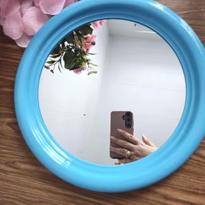 Op de afbeelding: Een ronde blauwe spiegel met een witte lijst. De spiegel reflecteert een persoon die een smartphone vasthoudt.