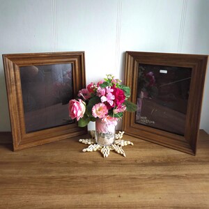 May include: Two wooden picture frames with a vase of pink and white flowers in the center. The frames are brown and have a simple design. The flowers are in a clear glass vase and are surrounded by seashells.