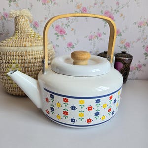 Peut inclure: Une bouilloire à thé en émail blanc ornée d'un motif répétitif de petites fleurs rouges, jaunes et bleues. La bouilloire possède une anse en bois clair et un bouton assorti sur le couvercle. Elle comprend un bec verseur et un corps arrondi.