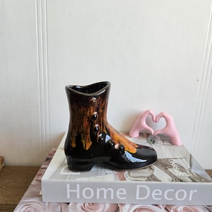 May include: A ceramic boot-shaped vase in glossy black and brown with button details. The vase sits atop two decorative books, one with "Home Decor" and the other with "Pink Rose" printed on the spines. A pink hand-shaped ornament is also present.