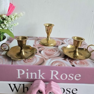 Peut inclure: Trois bougeoirs vintage en laiton. Deux ont une base en forme de soucoupe avec des poignées, et l'un est de forme classique. Ils sont disposés sur un livre avec les mots "Pink Rose" et un fond floral.