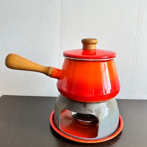 May include: A vintage fondue pot set with a vibrant orange gradient enamel pot, wooden handle and lid, and a gray and orange metal stand. The set is designed for tabletop cooking and serving.