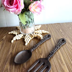 Puede incluir: Un juego de decoración de pared de hierro fundido en forma de cuchara y tenedor. La cuchara y el tenedor son marrones y tienen un aspecto rústico. Están colgados en una pared de madera.
