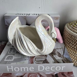 May include: A decorative, pearlescent white ceramic swan planter. The swan has a curved neck and an open basin. It rests on a stack of books with the words "Home Decor". A pink object is behind the swan.