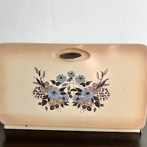 May include: A vintage beige bread bin with a floral design featuring blue and brown flowers. The bin has a brown plastic handle on the top.