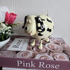Peut inclure: Une figurine de chien dalmatien crème avec des taches et des oreilles noires. Le chien a une expression souriante et se tient sur quatre pattes. La figurine est posée sur une pile de livres avec les titres "Rose" et "Pink Rose".
