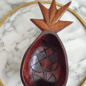 May include: A wooden pineapple-shaped bowl with a dark brown finish. The bowl has a carved pineapple design on the top and a carved pineapple design on the inside.