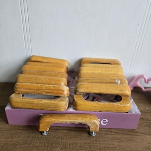 Może przedstawiać: Kolekcja drewnianych uchwytów do szafek w stylu vintage w różnych rozmiarach. Uchwyty są w kolorze jasnobrązowym z widocznymi śladami zużycia, co sugeruje wiek. Niektóre uchwyty są ułożone w stos, inne ułożone na fioletowym pudełku.