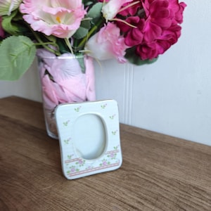 Peut inclure: Un cadre photo en céramique blanche avec un motif floral. Le cadre a un motif floral rose et vert et une ouverture ovale pour une photo.