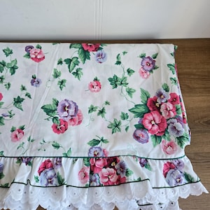 May include: White floral fabric featuring pink, purple, and green pansies and vines. The fabric has a ruffled edge with white eyelet lace and a green trim. The fabric is folded on a wooden surface.
