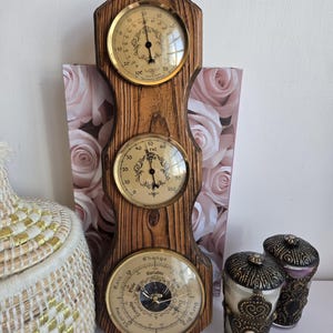 May include: A wooden weather station with three circular gauges. The top gauge displays temperature in Fahrenheit, the middle gauge shows relative humidity, and the bottom gauge is a barometer. Two decorative jars with ornate lids are also visible.