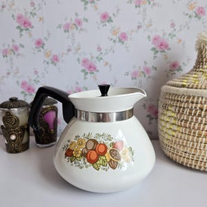 May include: A white teapot with a black handle and a small black lid knob. The body features a colourful botanical print of nuts, leaves, and yellow flowers. A silver-toned metal band encircles the upper portion of the teapot below the lid.