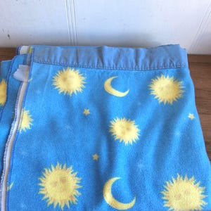 May include: A blue fleece blanket featuring a repeating pattern of yellow suns, crescent moons, and stars. The blanket has a light blue border and is folded. The blanket is made of a soft material.