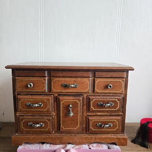 Peut inclure: Un coffret à bijoux en bois vintage avec plusieurs tiroirs et des poignées en métal ouvragées. La boîte est de couleur marron chaud avec des accents dorés. Le mot "Pose" est visible sur une boîte rose devant le coffret.