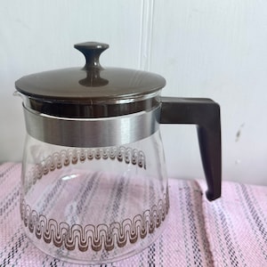 May include: A brown and clear glass coffee pot with a brown lid and a silver band around the middle. The pot has a brown geometric pattern around the bottom.