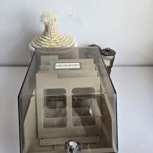 Peut inclure: Un classeur de cartes Microport beige avec un couvercle en plastique transparent et une serrure avec des clés. Le classeur comporte plusieurs fentes pour les cartes. Le texte "MICROPORT" est visible sur une étiquette. Un panier décoratif tissé est en arrière-plan.