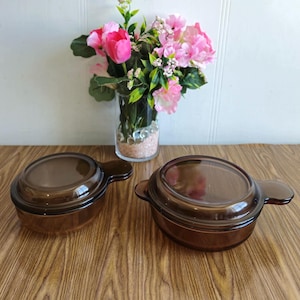 Op de afbeelding: Twee bruine glazen ovenschaaltjes met deksels en handvatten. De schaaltjes staan op een houten oppervlak.