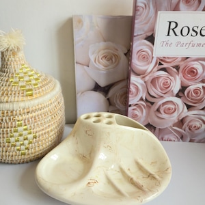 May include: A cream-coloured, seashell-shaped ceramic toothbrush holder with brown marbling. The holder has six holes for toothbrushes and a curved design. A woven basket with gold accents and a book titled "Rose" are in the background.
