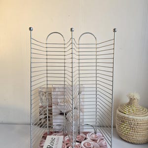 Puede incluir: Un estante de alambre de metal plateado con una base floral rosa, que muestra el texto "Pink Rose". El estante tiene un diseño decorativo con tapas redondeadas y contiene un libro con el texto "Rose The Perfumes."