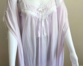 Puede incluir: Un conjunto de camisón y bata de color lavanda claro. El camisón presenta tirantes finos, un borde de encaje blanco a lo largo del escote en V y un pequeño lazo de cinta blanca en el centro. La bata tiene mangas cortas y cierre de botones.