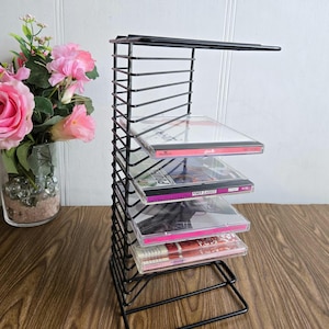 Op de afbeelding: Zwarte metalen CD-rek met vijf rijen CD's. Het rek staat op een houten oppervlak.