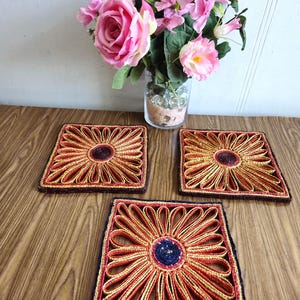 May include: Three woven square coasters featuring a red, gold and black floral design. The coasters are made from natural materials and have a rustic appearance.
