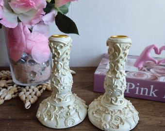 May include: A pair of white candle holders with intricate floral designs and gold accents. The candle holders are on a wooden surface.