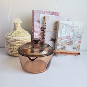 Op de afbeelding: Een heldere, amberkleurige glazen steelpan met een deksel en handvat. Een geweven mand met een deksel en decoratieve boeken met bloemenkaften zijn ook aanwezig. De boeken heten "Pink Roses", "White Roses" en "Home Decor".
