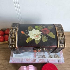 May include: A black wooden chest with a floral design on the lid. The chest has gold trim and a metal clasp. It is sitting on top of a stack of books. A pink object and a red fez are in the foreground.