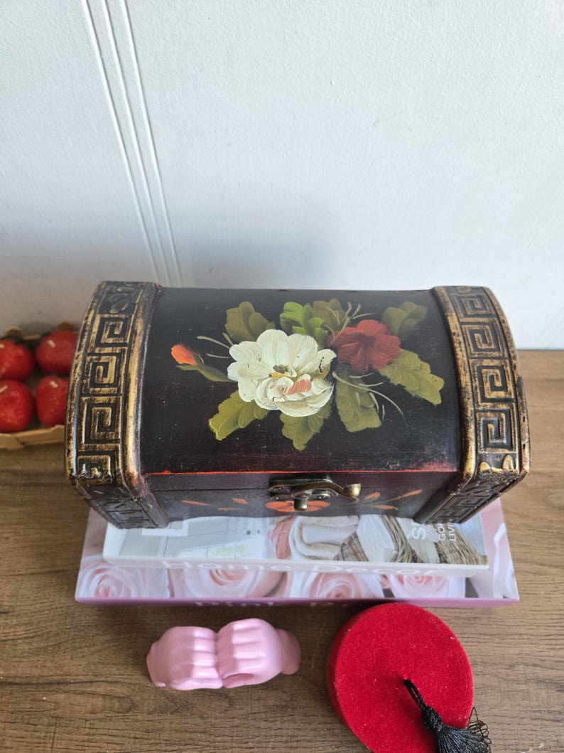 May include: A black wooden chest with a floral design on the lid. The chest has gold trim and a metal clasp. It is sitting on top of a stack of books. A pink object and a red fez are in the foreground.