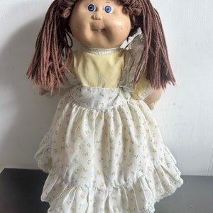 May include: A vintage Cabbage Patch Kid doll with brown yarn hair, blue eyes, and a yellow shirt with a white floral print dress.