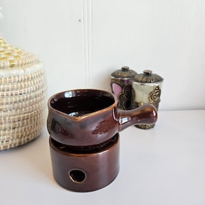 Peut inclure: Un chauffe-huile en céramique marron avec une poignée, posé sur une base cylindrique avec un trou. Deux pots décoratifs avec des couvercles ornés sont en arrière-plan. Le chauffe-huile a une finition brillante.
