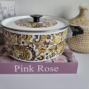 Peut inclure: Une casserole blanche et en acier inoxydable avec un couvercle. Le pot présente un motif cachemire dans les tons marron et jaune. Les poignées et le bouton du couvercle sont noirs. Le pot est posé sur un livre qui dit "Pink Rose".