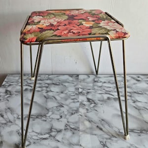 May include: A vintage vanity stool with a floral patterned seat. The seat features a mix of red, pink, and green flowers. The stool has a metal frame with four angled legs and measures approximately 15 inches tall.