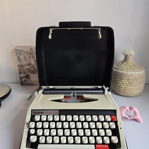 Puede incluir: Máquina de escribir Brother Deluxe color crema con teclas negras y una funda negra. La máquina de escribir tiene una tecla de tabulación roja y una tecla de espaciado de repetición. Una cesta decorativa tejida y una escultura rosa en forma de mano están al lado.